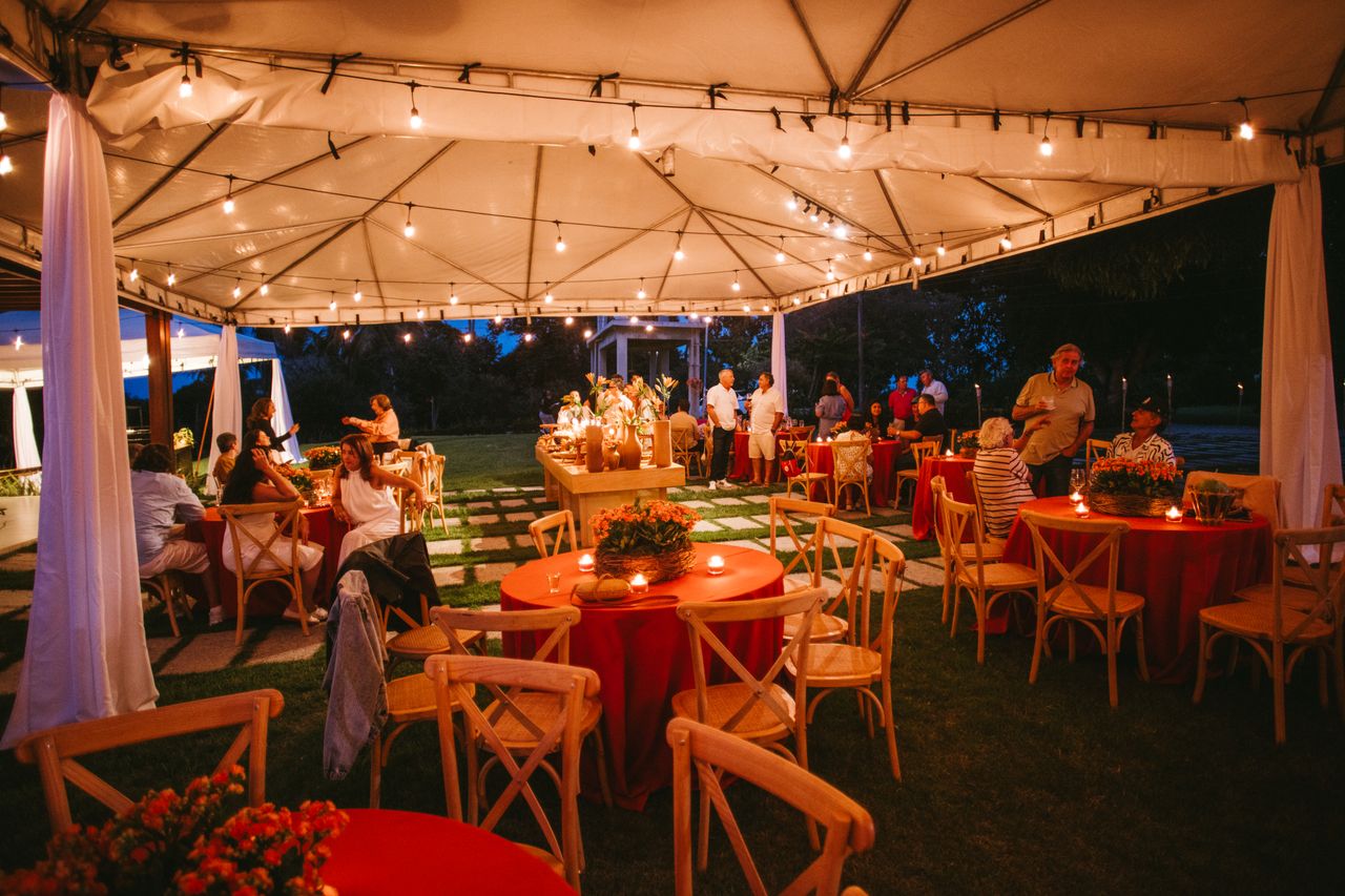 gallery-01 Charming outdoor evening event setup with string lights, red tablecloths, and wooden chairs under a canopy.